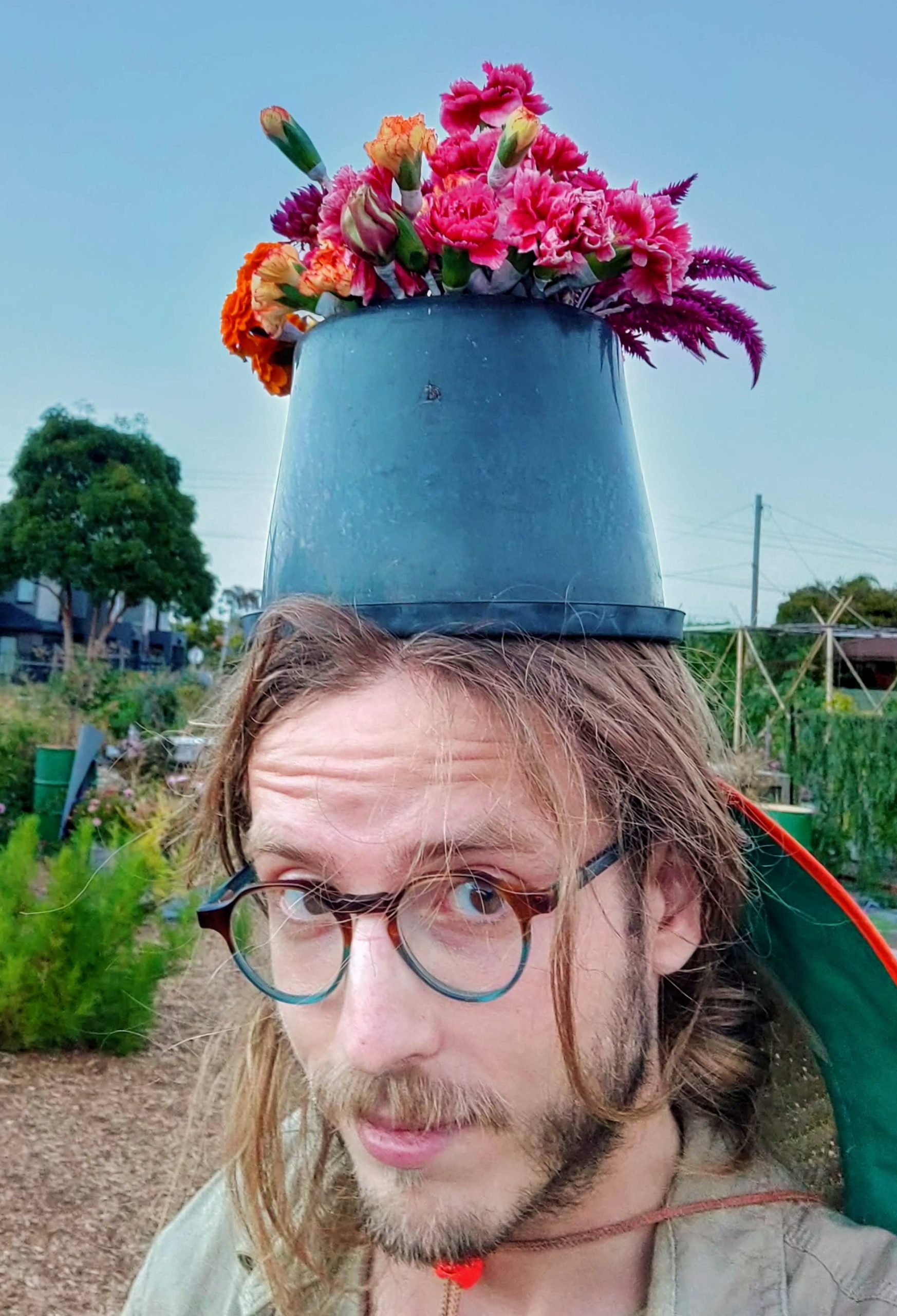 A photograph of Ollie wearing a flowerpot, on site at the build and installation of a bamboo frame for a wedding in a local community garden.